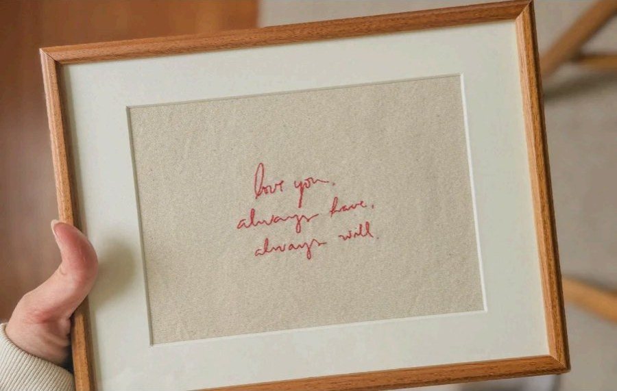 A short handwritten note stitched in red thread, framed behind a mat, held in one hand